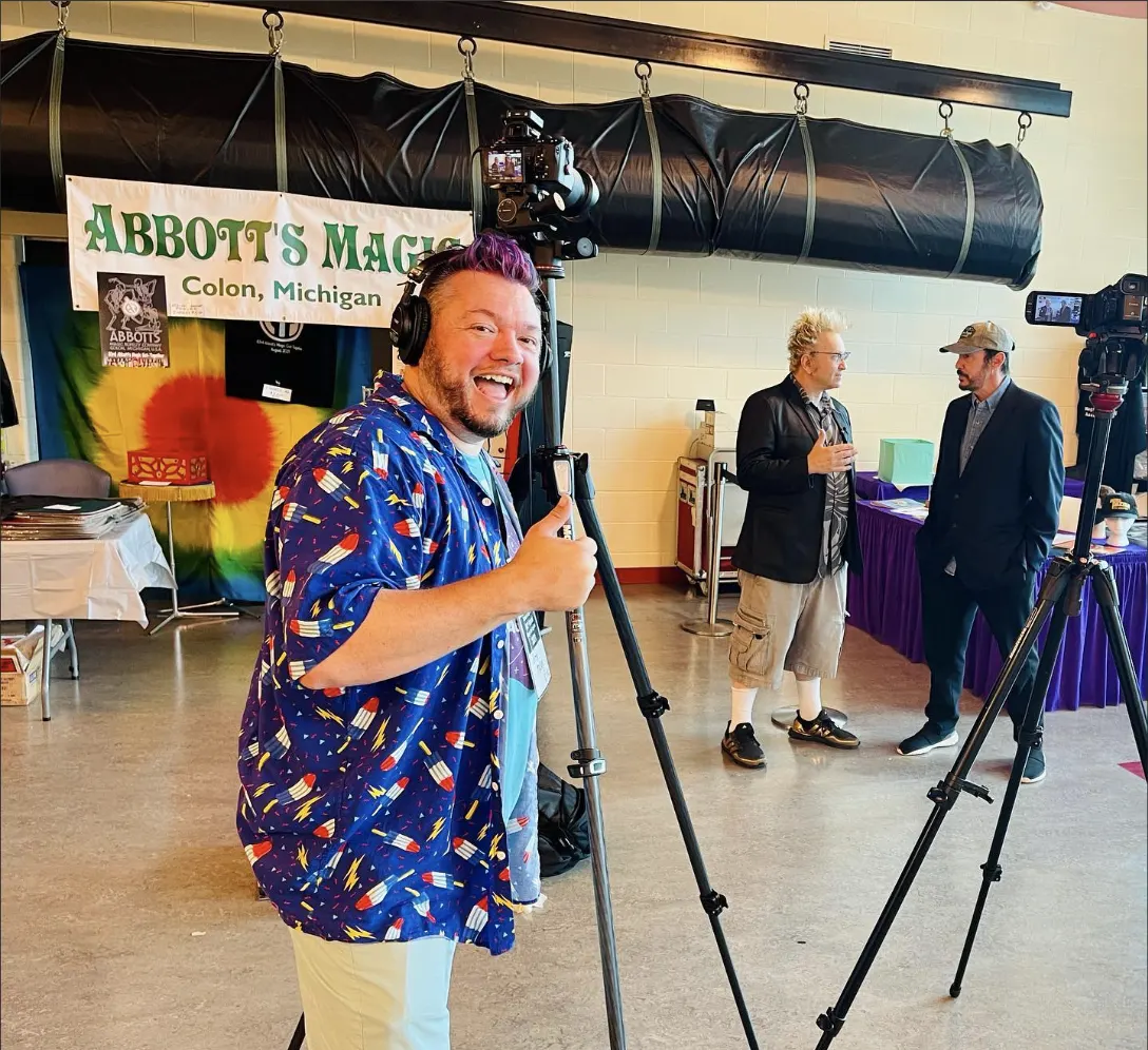 Greg Mulvey grinning behind a camera on a tripod at the Abbott's Magic Get Together in Colon, Michigan